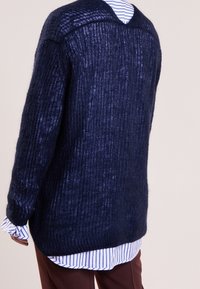 Granatowy sweter dzianinowy z żebrowaną fakturą, z szerokim okrągłym dekoltem i luźnym krojem. Noszony na niebieskiej i białej koszuli w paski, z długimi rękawami.