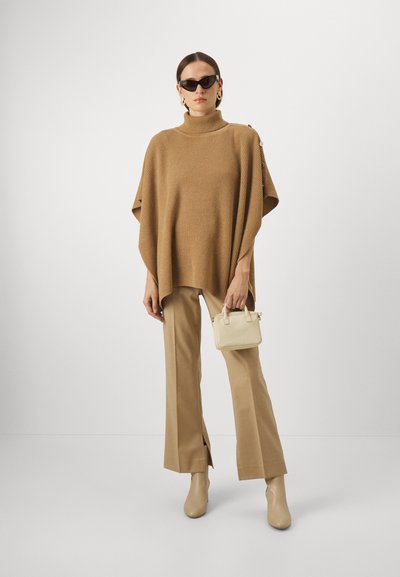 Kvinne iført en kamelfarget ribbet poncho, matchende vide bukser, beige støvler, svarte solbriller, gull hoops-øreringer og holder en liten kremfarget håndveske.