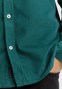 Teal kordfløjl skjorte med lodret ribbing, en spids krave og hvide knapper, samt et mærke ved kanten. Sorte jeans synlige nedenfor.