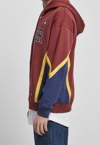 Sweat à capuche bordeaux avec un design diagonal jaune et bleu, tissu doux, poche kangourou, et logo brodé sur la poitrine.