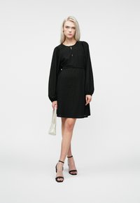 Γυναίκα που στέκεται σε ένα μαύρο μακρυμάνικο φόρεμα με ζώνη, κρατώντας μια μικρή λευκή τσάντα, φορώντας μαύρες ανοιχτές γόβες σε μια απλή φόντο.