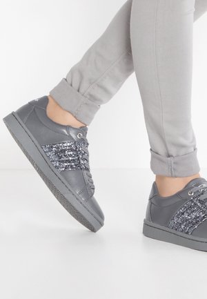 Personne portant un pantalon gris clair retroussé et des baskets grises avec des rayures noires pailletées, debout sur un sol blanc.