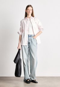 Svijetlo ružičasta košulja dugih rukava preko bijelog topa, uparena s plavim širokim trapericama i crnim cipelama, drži crnu torbu. Jednostavan dizajn i materijali.