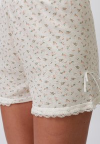Pantalones cortos con estampado floral sobre un fondo crema, que presentan pequeñas flores rosas y verdes, con un borde de encaje en el dobladillo y un lazo decorativo en el lado.