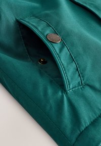 Detailná záber na tyrkysové látkové vrecko so zapínaním na patent, zobrazujúce detaily šitia a kovový povrch patentu.