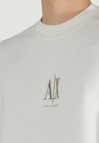 Weißes T-Shirt mit kurzen Ärmeln aus weichem Stoff, das ein braunes gesticktes "AX"-Logo mit dem Text "ARMANI EXCHANGE" darunter zeigt, runder Halsausschnitt.