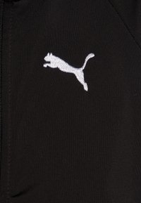 Tissu noir avec une texture lisse présentant un logo Puma brodé en blanc d'un animal sautant. Détails de fermeture éclair également visibles.