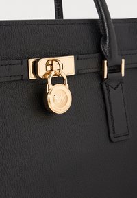 Zwarte leren handtas met een gouden slot met de initialen "MK", een gladde textuur en een gestructureerd ontwerp met stevige handvatten.