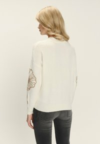 Blonde Frau, die seitlich gedreht ist und einen cremefarbenen Pullover mit beige bestickten Blumen an den Ärmeln und dunkelgrauen Jeans trägt.