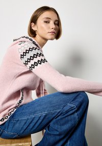 Różowa dziergana bluza z kapturem i sznurkiem, z czarnym geometrzycznym wzorem na ramionach. Połączona z niebieskimi dżinsami w kształcie dzwonów. Widoczna miękka tekstura.