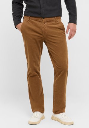Braune Cord-Hosen mit strukturierter Oberfläche, schmaler Schnitt, seitlichen Taschen und Knopfverschluss. Kombiniert mit weißen Schnürsneakern.