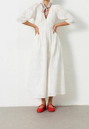 Femme portant une robe midi blanche brodée, un foulard à motifs autour du cou et des ballerines rouges, posant les mains sur les hanches contre un mur blanc.
