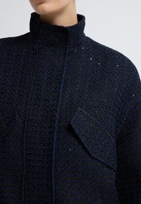 Granatowa kurtka teksturowana z wysokim kołnierzem, z plecionym wzorem oraz dwiema kieszeniami naszytymi. Iskrzące akcenty podkreślają wizualne zainteresowanie materiału.