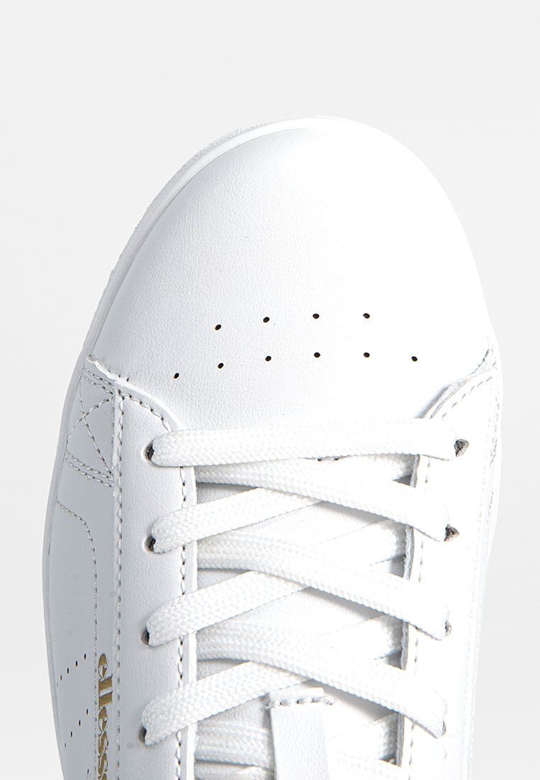 Weiße Ledersneaker mit durchlöchertem Obermaterial, flachen Schnürsenkeln und einem strukturierten Design. Goldene Markenapplikation an der Seite. Abgerundete Zehenpartie.