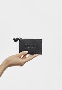 Portafoglio in pelle nera testurizzata con logo "TOUS" in rilievo, chiusura con zip e accento in silicone nero, tenuto in una mano su uno sfondo bianco.
