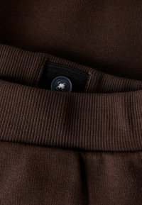 Brunt bomullsstoff med ribbet tekstur og en flat linning. Har en plastknapp og sydd detaljer for lukking.