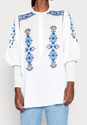 Hvid bluse med blå blomsterbroderi foran, på skuldre og ærmer, med knaplukning og ballonærmer med manchet.