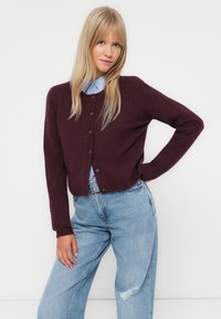 Gilet en tricot bourgogne avec fermeture à boutons, manches longues et coupe droite. Associé à une chemise bleu clair et à un jean bleu décoloré.
