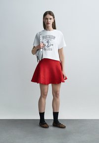 Γυναίκα που φοράει λευκό μπλουζάκι με το κείμενο "Paris Amour", κόκκινη πλισέ φούστα, μαύρες κάλτσες, παπούτσια με λεοπάρ σχέδιο και κρατάει λευκή τσάντα με φούντες.