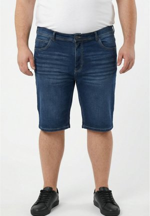 Mann mit blauen Jeansshorts und schwarzen Sneakers steht vor einem schlichten weißen Hintergrund.