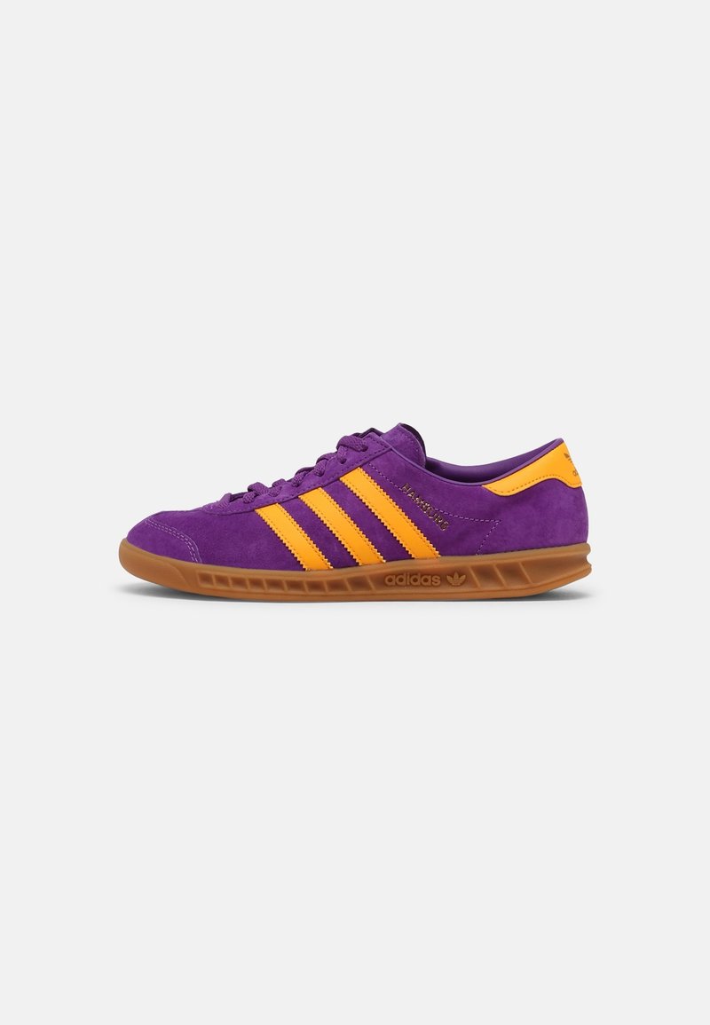Purple suede sneaker with three orange stripes, brown rubber sole, rounded toe, and padded collar. Classic athletic design and textured surface.