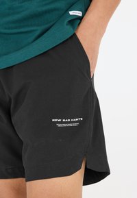 Svarta shorts i lätt tyg, med en sidoklyvning och vit tryckt text längs fållen. Matchas med en teal-färgad skjorta.