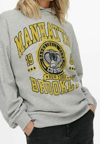 Felpa grigia con una grande stampa grafica gialla e nera che riporta "MANHATTAN BROOKLYN 1988" e un motivo di pallacanestro. Materiale in cotone, vestibilità rilassata.