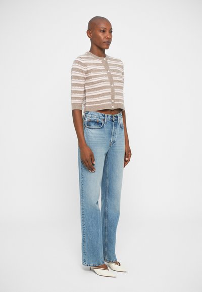 Cardigan en tricot à rayures beige et blanches avec des manches courtes, fermeture à boutons, associé à un jean taille haute bleu clair à jambes larges et des escarpins blancs pointus.