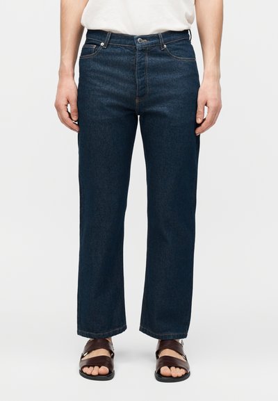 Personne portant un jean bleu foncé à jambes droites et des sandales marron ouvertes à l'avant, debout devant un fond blanc uni.