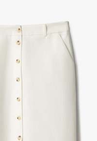 Jupe blanche avec une fermeture à boutons sur le devant composée de six boutons ronds couleur crème, taille plate et poches latérales. Texture lisse.