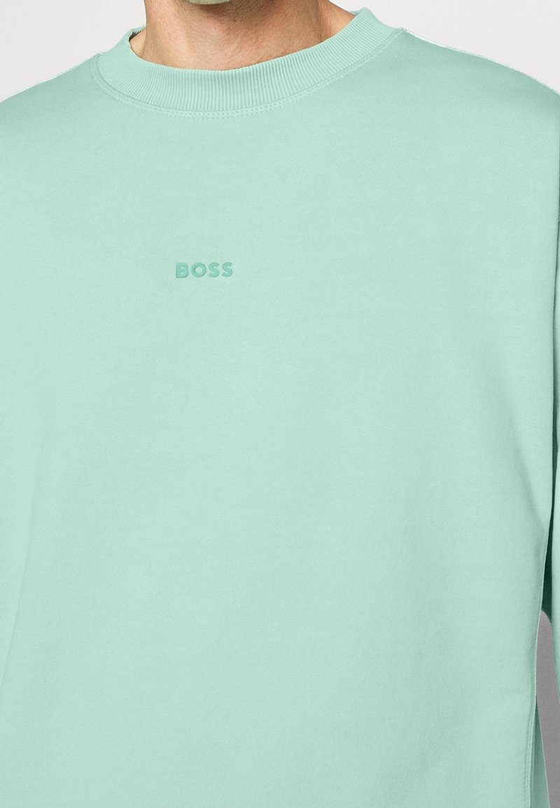 Homme portant un sweat-shirt col rond vert menthe avec le logo "BOSS" en relief centré sur la poitrine.