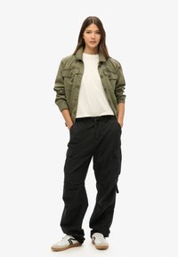 Olijfgroene jas met twee borstzakken, wit T-shirt, zwarte cargobroek en witte sneakers met zwarte accenten. Casual pasvorm en ontspannen ontwerp.