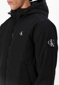 Osoba ubrana w czarną kurtkę z kapturem zapinaną na zamek, z białym logo "cK" na piersi i naszywką na rękawie, stojąca na jednolitym tle.