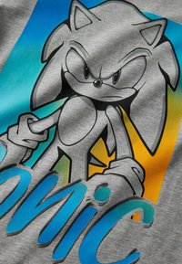Szara tkanina z nadrukowaną postacią z kreskówki o kolczastych włosach, na tle gradientu w kolorze niebieskim i żółtym, z napisem „Sonic” w pogrubionym niebieskim tekście.