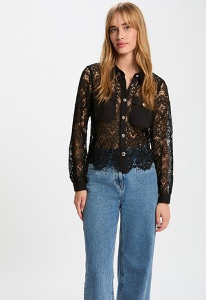 Ung kvinde med langt blondt hår iført en sort blondebluse med knapper og højtaljede blå jeans mod en hvid baggrund.