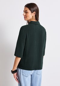 Camicia corta a maniche con colletto di un verde scuro e testa ruvida, abbinata a jeans in denim azzurro chiaro. La camicia presenta sottili righe verticali.