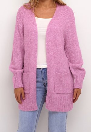 Femme portant un cardigan duveteux rose clair ouvert sur le devant avec deux poches avant, par-dessus un haut blanc et un jean bleu clair.