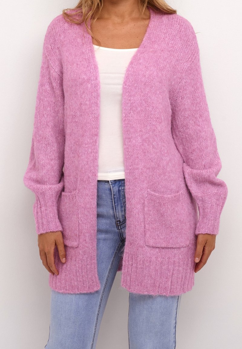 Femme portant un cardigan duveteux rose clair ouvert sur le devant avec deux poches avant, par-dessus un haut blanc et un jean bleu clair.