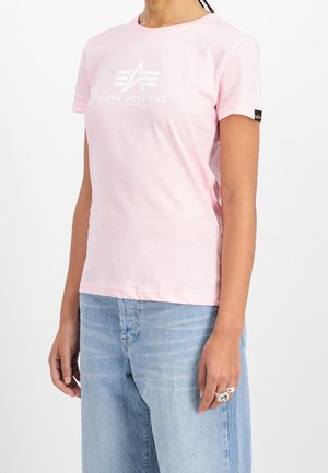 Kobieta w jasno różowej koszulce Alpha Industries i jasnoniebieskich dżinsach, pokazująca logo i metkę na rękawie, stoi na białym tle.