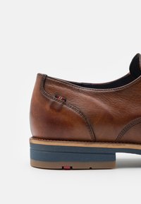 Zapato de cuero marrón con puntera redondeada, suela en contraste azul y marrón, y un pequeño acento a rayas cerca del talón. Textura suave.