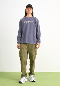 Lilla langærmet sweatshirt med limegrønne accenter og grafisk tekst, parret med olivengrønne cargo-bukser og hvide sneakers.