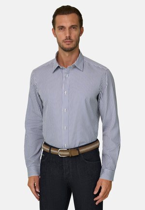 Homme portant une chemise à manches longues à rayures verticales bleues et blanches et un pantalon foncé avec une ceinture beige tissée.
