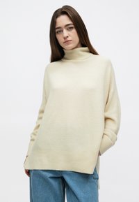 Kremo spalvos golfas, laisvo silueto, tekstūruoto mezginio audinys, šoninės skeltukės ir nukabintos pečiai, derinamas su šviesiai mėlynais džinsais.