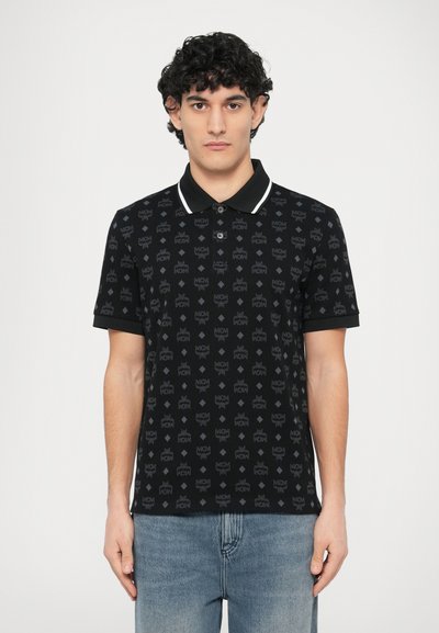 Polo noir avec un motif subtil de logos en all-over, col blanc contrastant, manches courtes et patte de boutonnage. Porté avec un jean bleu clair.