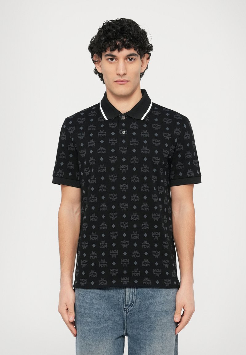 Polo preta com padrão sutil de logótipos em todo o tecido, gola branca contrastante, mangas curtas e uma pala de botões. Combinada com jeans azul claro.