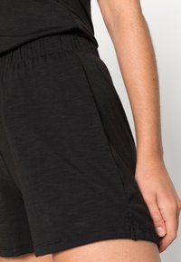 Calções pretos feitos de um tecido leve e texturizado. Apresentam uma cintura elástica e bolsos laterais. A bainha tem um discreto detalhe de costura.