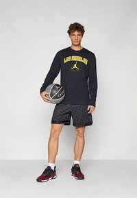 Fiatal férfi fekete "Los Angeles" hosszú ujjú pólóban, mintás rövidnadrágban és piros-fekete tornacipőben, aki kosárlabdát tart egy egyszínű szürke háttér előtt.