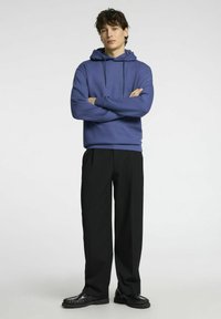 Felpa blu con cappuccio e coulisse regolabili, abbinata a pantaloni neri e scarpe nere. Tessuto morbido, vestibilità rilassata e silhouette tradizionali.
