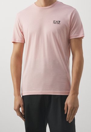 Mężczyzna ubrany w jasnoróżowy krótki rękaw t-shirt z logo EA7 Emporio Armani na lewej piersi i czarne spodnie.
