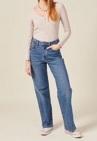 Maglietta a maniche lunghe color beige chiaro a coste con scollatura a barca e dettaglio dei bottoni, abbinata a jeans blu a vita alta e scarpe da ginnastica bianche.